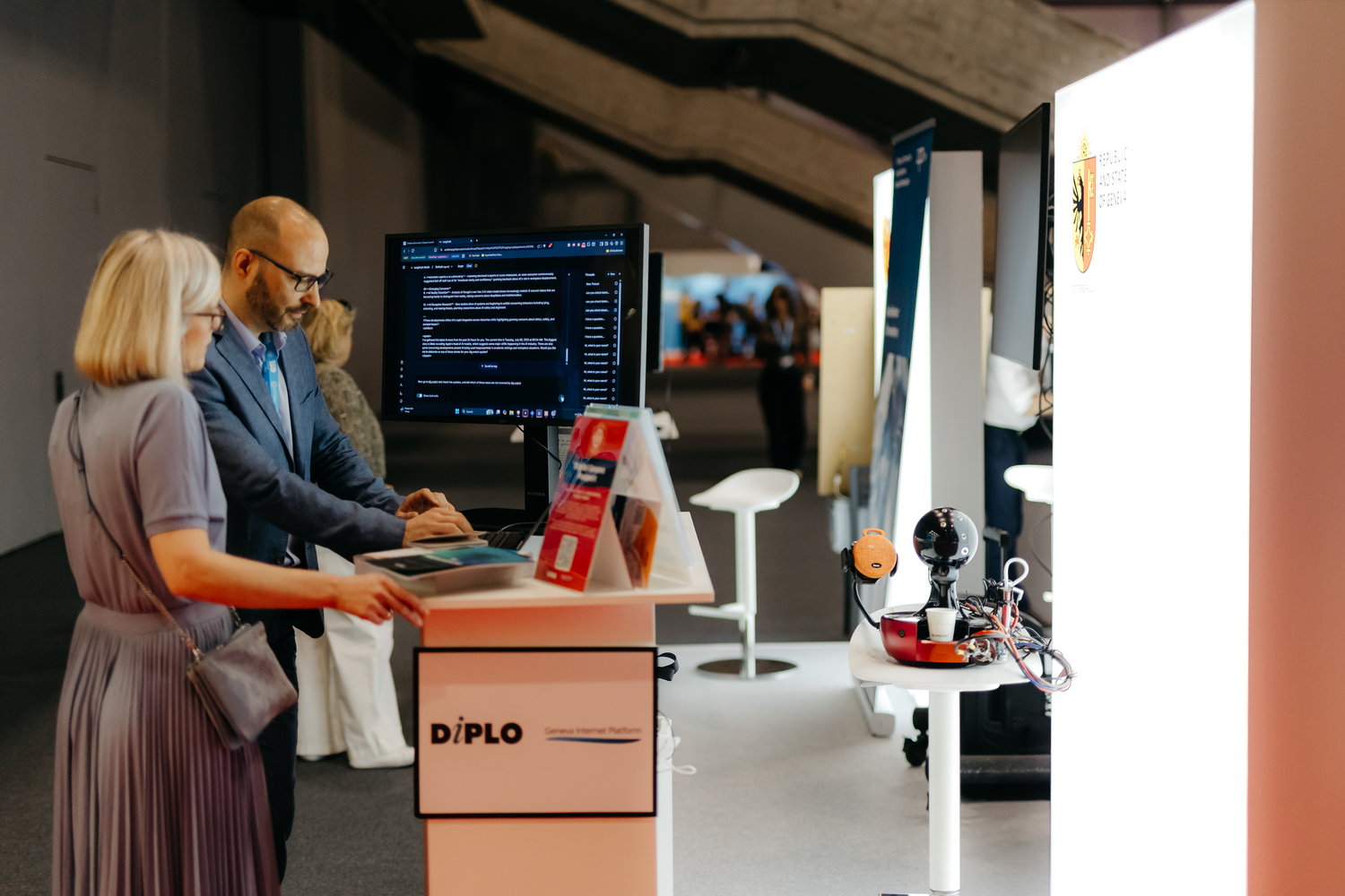 Diplo/GIP at AI for Good Global Summit 6 Furniture, Table, Desk, Adult, Female, Person, Woman, Electronics, Screen, Computer Hardware, Hardware, Monitor, Box, Face, Head, Computer, Pc, Indoors, Accessories, Bag, Handbag, Clothing, Coat